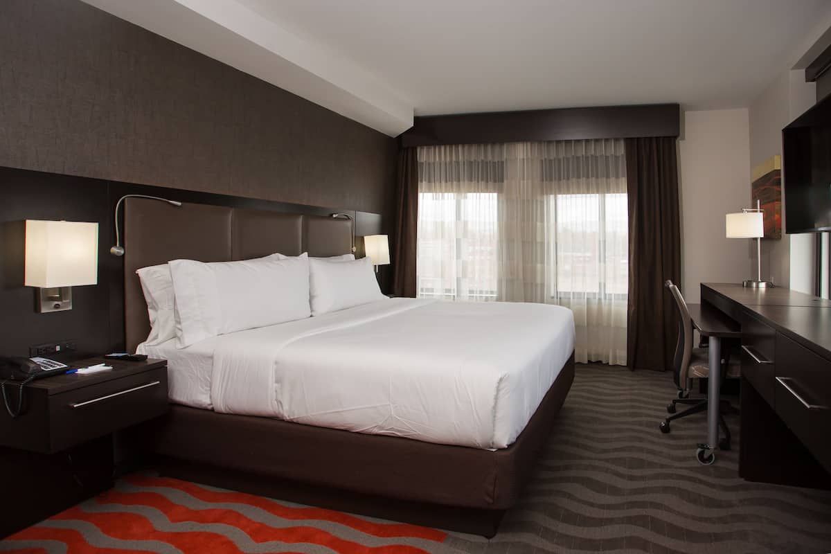 A hotel room with a large bed, a desk, and a chair. There are two bedside lamps and a window with curtains.