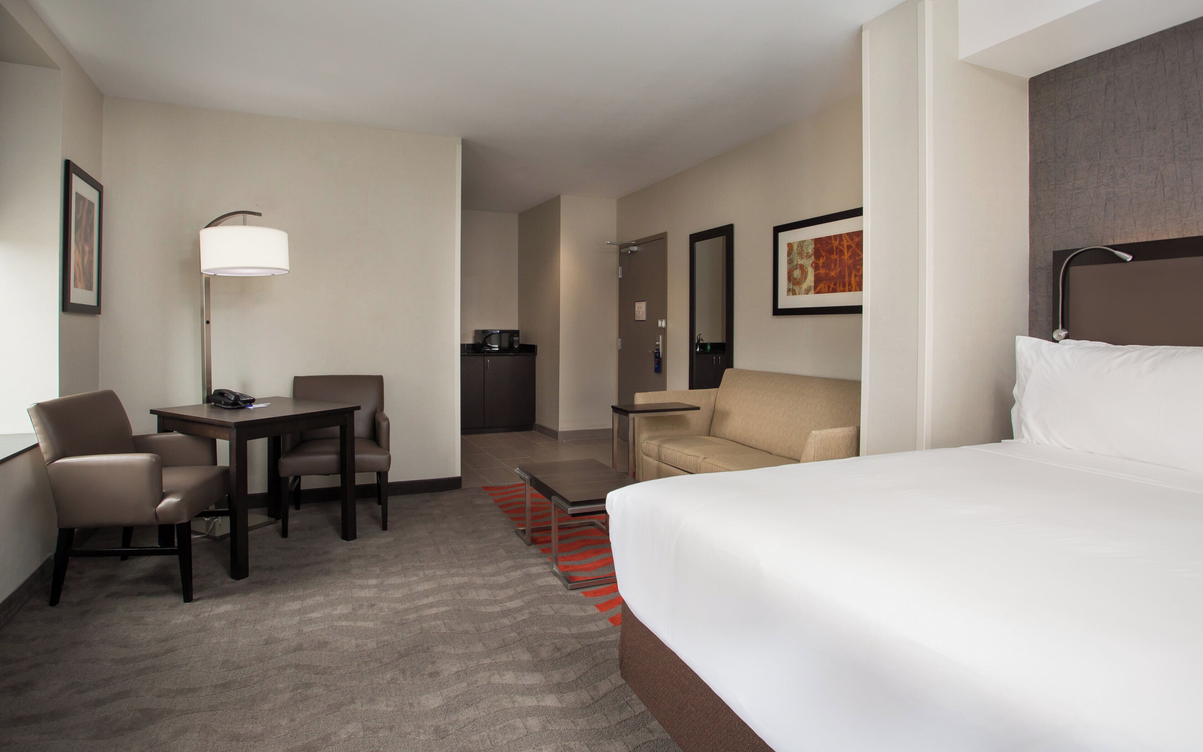 A hotel room with a bed, a desk, a chair, a sofa, a lamp, and a small kitchenette.
