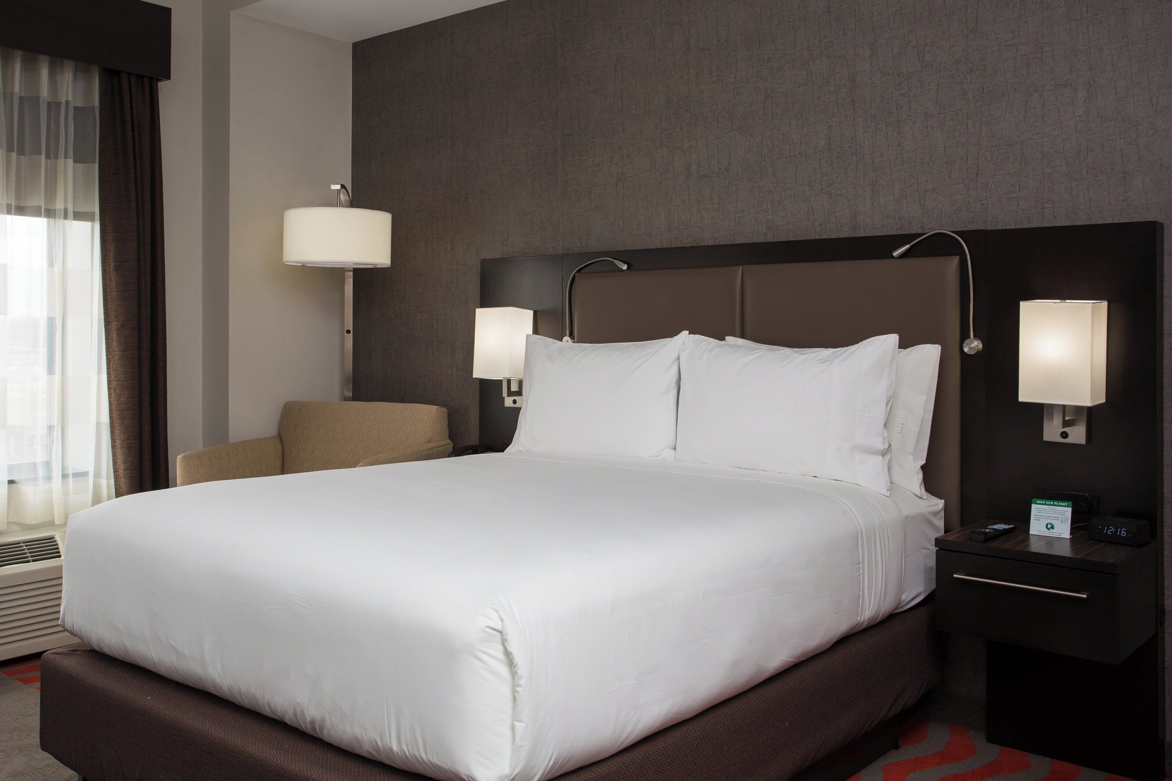 Habitación de hotel con una cama grande, dos lámparas de noche, una silla y una mesita de noche con un reloj.