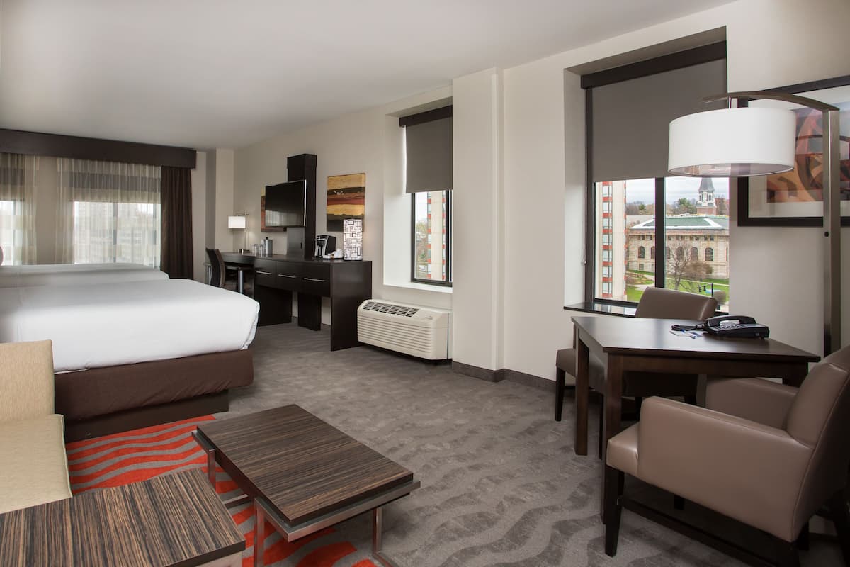 A hotel room with a bed, a desk, a chair, a small table, and a window with a view of a building.