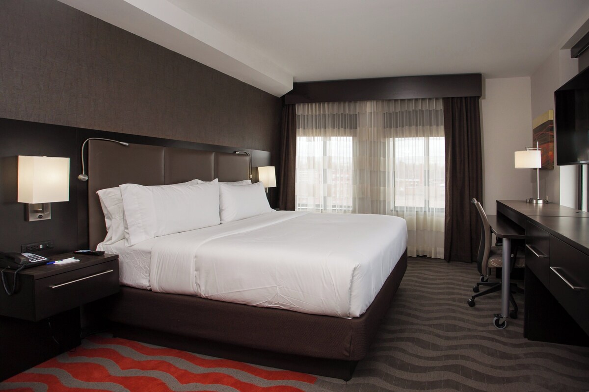 A hotel room with a large bed, bedside tables with lamps, a desk with a chair, and a large window with curtains.