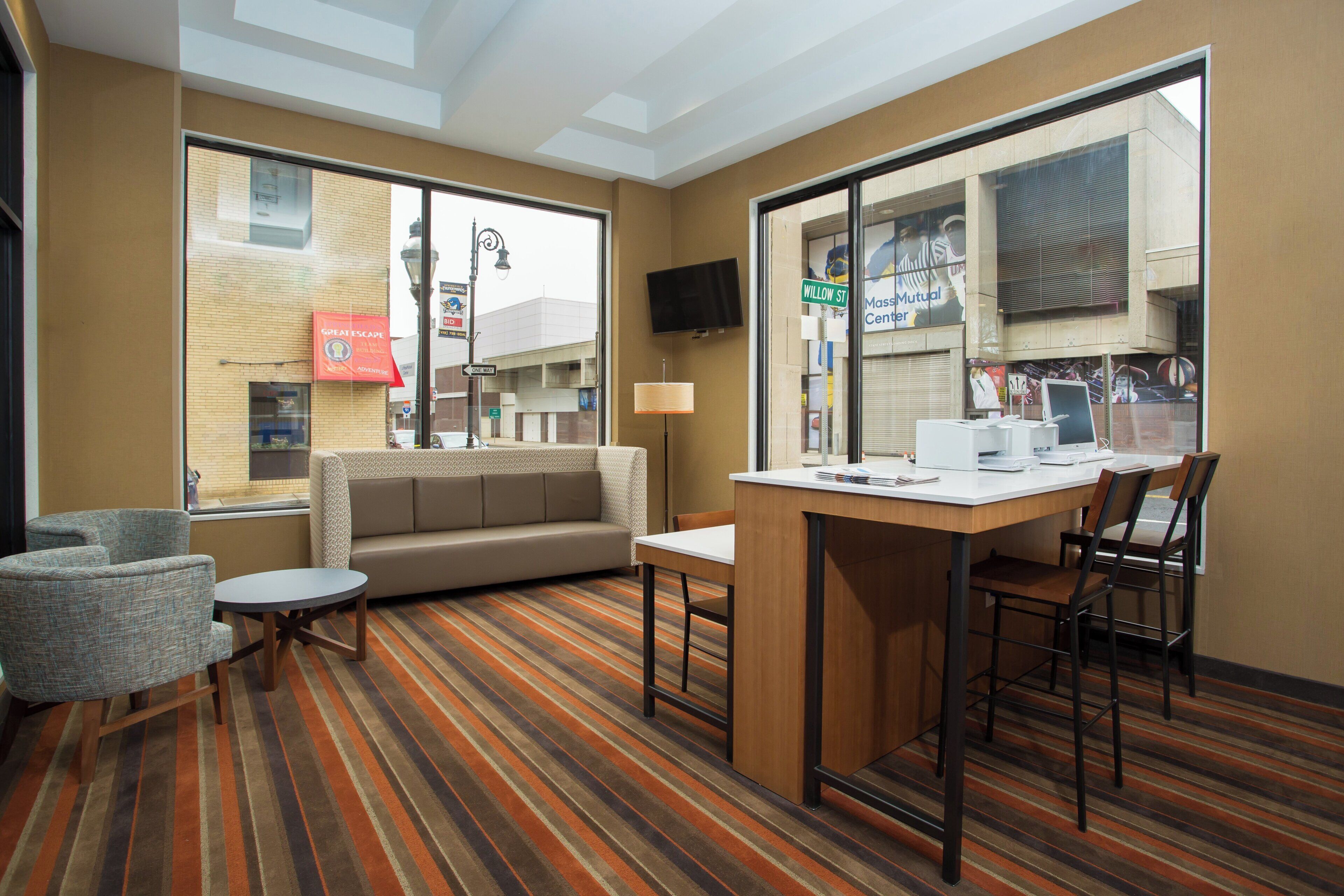 A hotel room with a sofa, a small table, a chair, a TV, and a window with a view of the street.