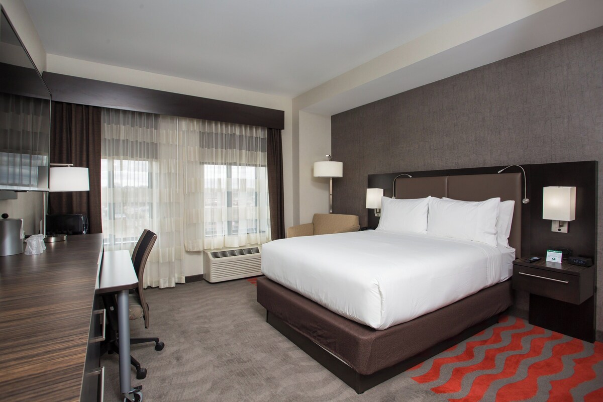 A hotel room with a large bed, a desk, a chair, a TV, and a window with curtains.