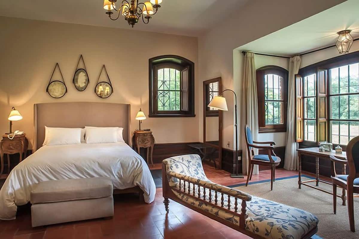 A bedroom with a large bed, bedside tables, a chair, and a desk with a lamp. There are three wall-mounted light fixtures and a window with curtains.