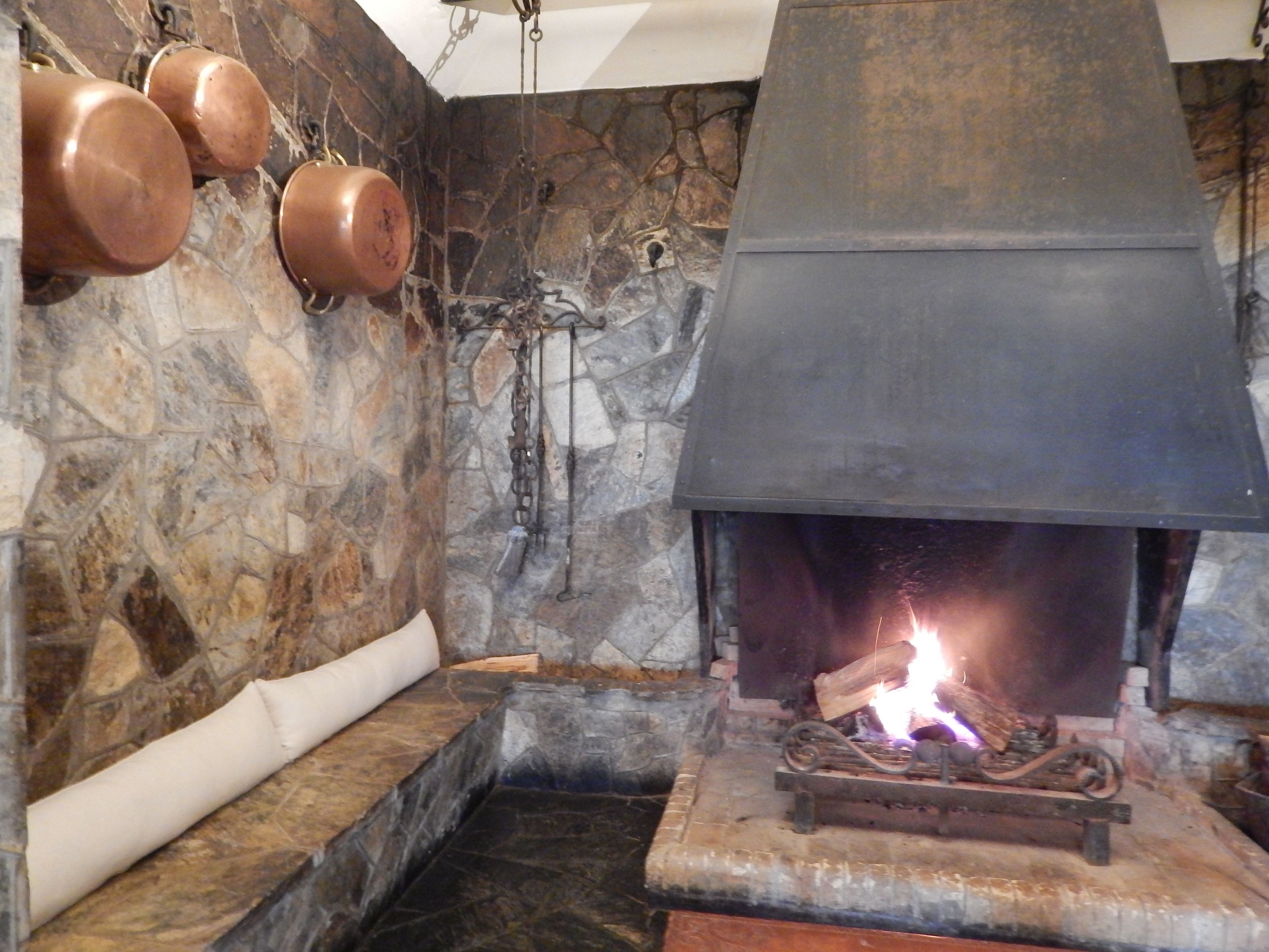 A stone-walled room with a fireplace, a stone bench, and hanging copper pots.