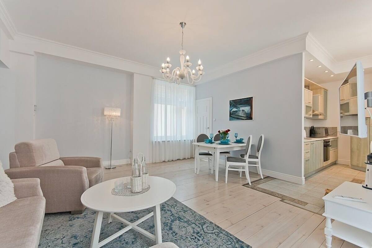 A modern living room with a dining area, a chandelier, and a kitchen in the background.