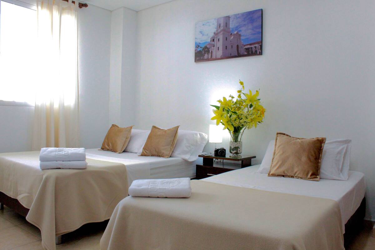 A hotel room with two beds, each with a white bedspread and beige pillows. There is a vase with yellow flowers on a nightstand between the beds.