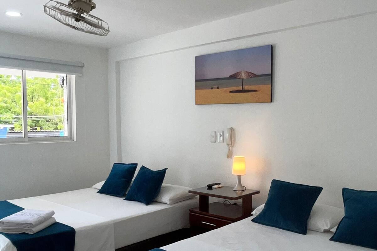 A hotel room with two beds, a window, a painting, a bedside table with a lamp, and a ceiling fan.