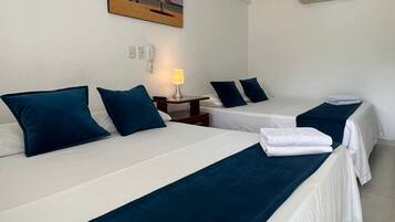 A hotel room with two beds, each with a white mattress and a dark blue bedspread. There is a painting of a beach scene on the wall above the beds, and a wall-mounted air conditioner to the right.