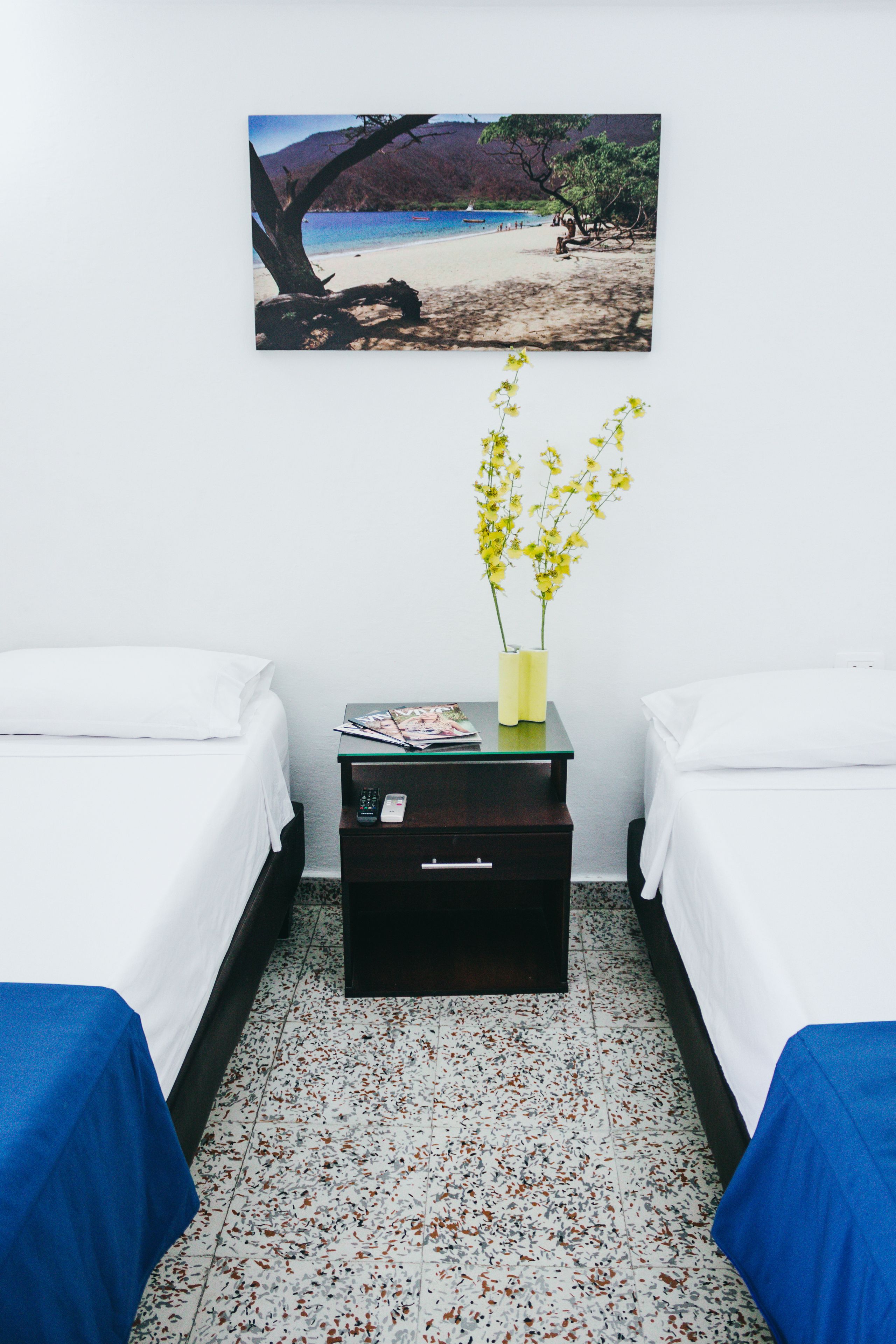 Ein Hotelzimmer mit zwei Betten, einem Nachttisch mit einer Vase gelber Blumen und einem Strandbild an der Wand.