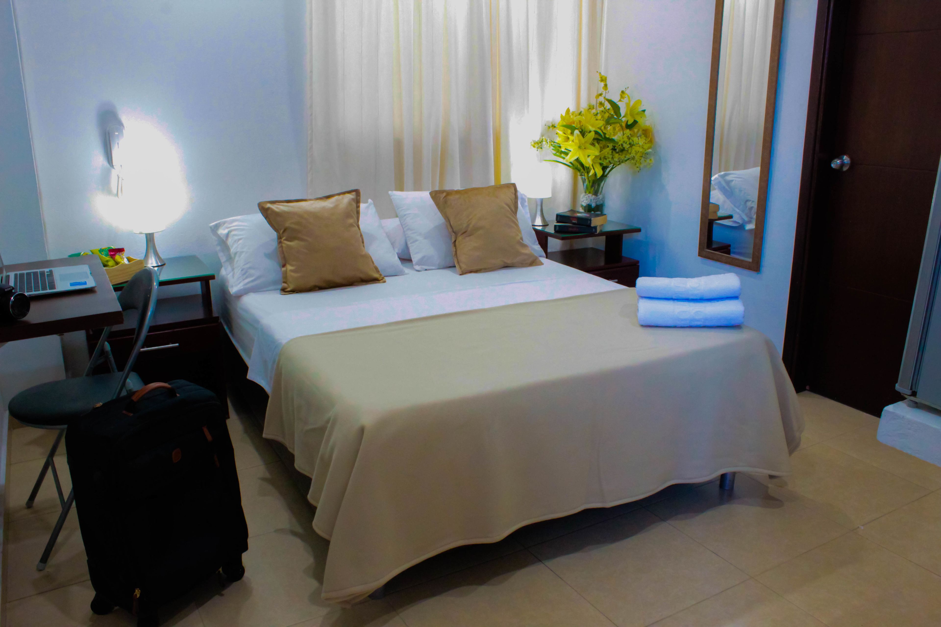 A hotel room with a bed, a desk with a laptop, a suitcase, a lamp, and a mirror.