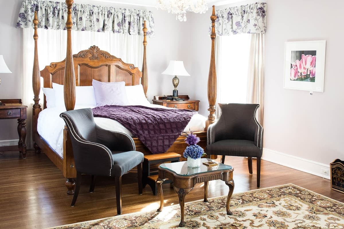 A four-poster bed with a wooden headboard, two bedside tables with lamps, a small table with a vase, two upholstered chairs, and a patterned rug on a wooden floor.