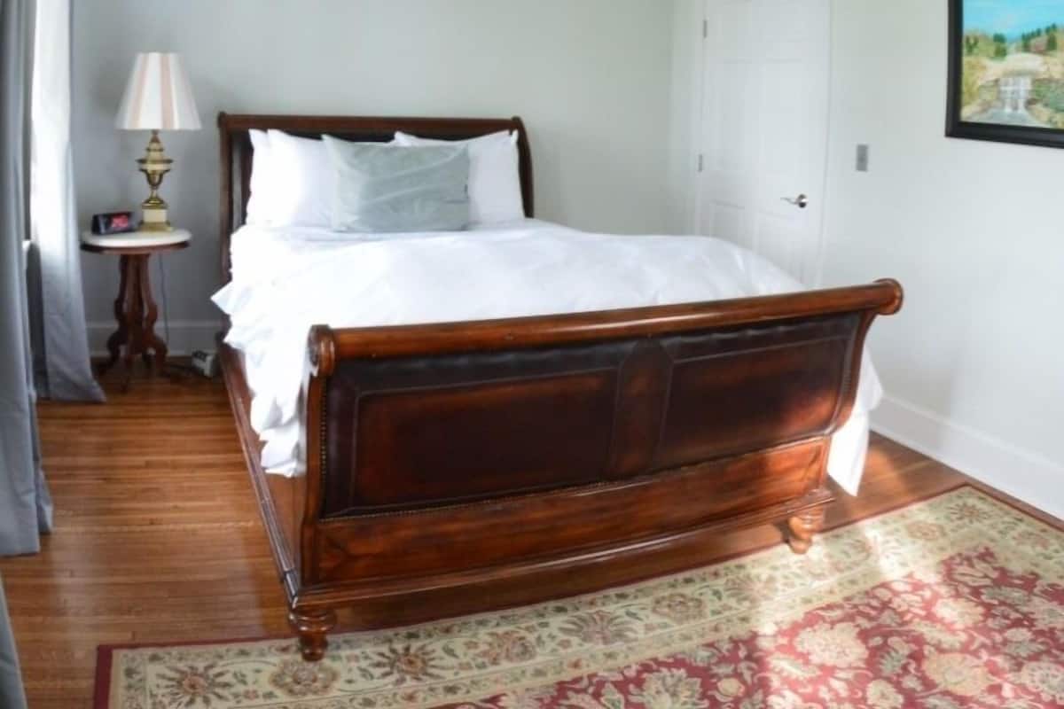 A neatly made bed with a wooden headboard in a room with a patterned rug, a small table with a lamp, and a door in the background.