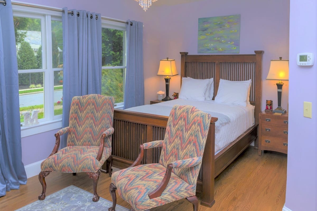 A bedroom with a bed, two upholstered chairs, a chandelier, a painting, and a thermostat.