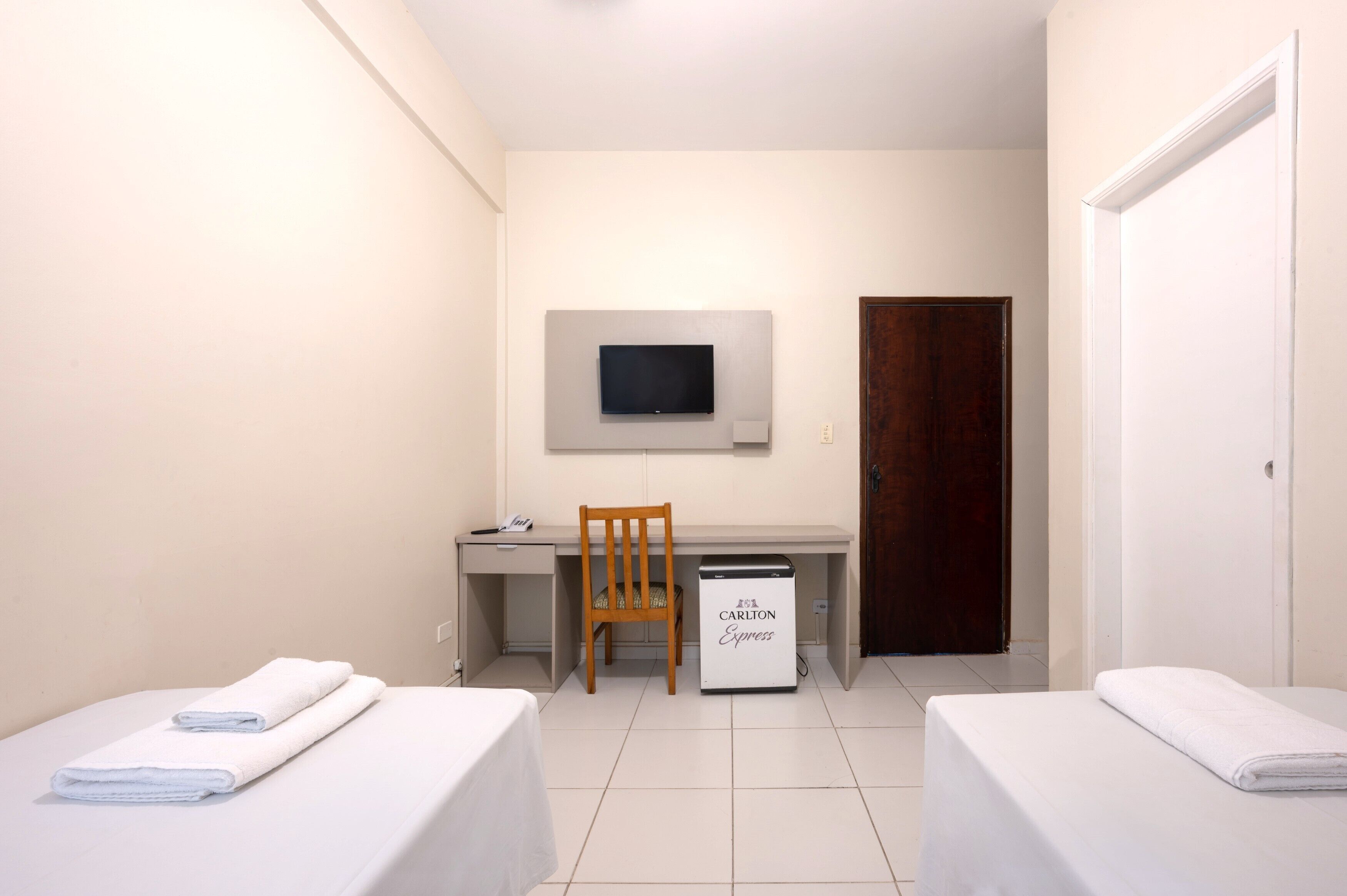 A small, clean room with two beds, a desk, a chair, a TV mounted on the wall, and a minibar.