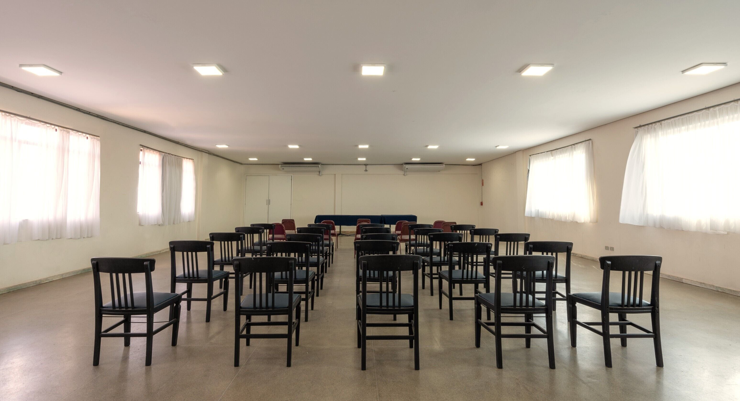 A conference room with rows of chairs facing a stage, white walls, and large windows with curtains.