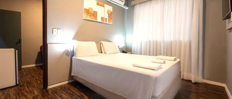 A hotel room with a large bed, a flat-screen TV, an air conditioning unit, and a window with curtains.