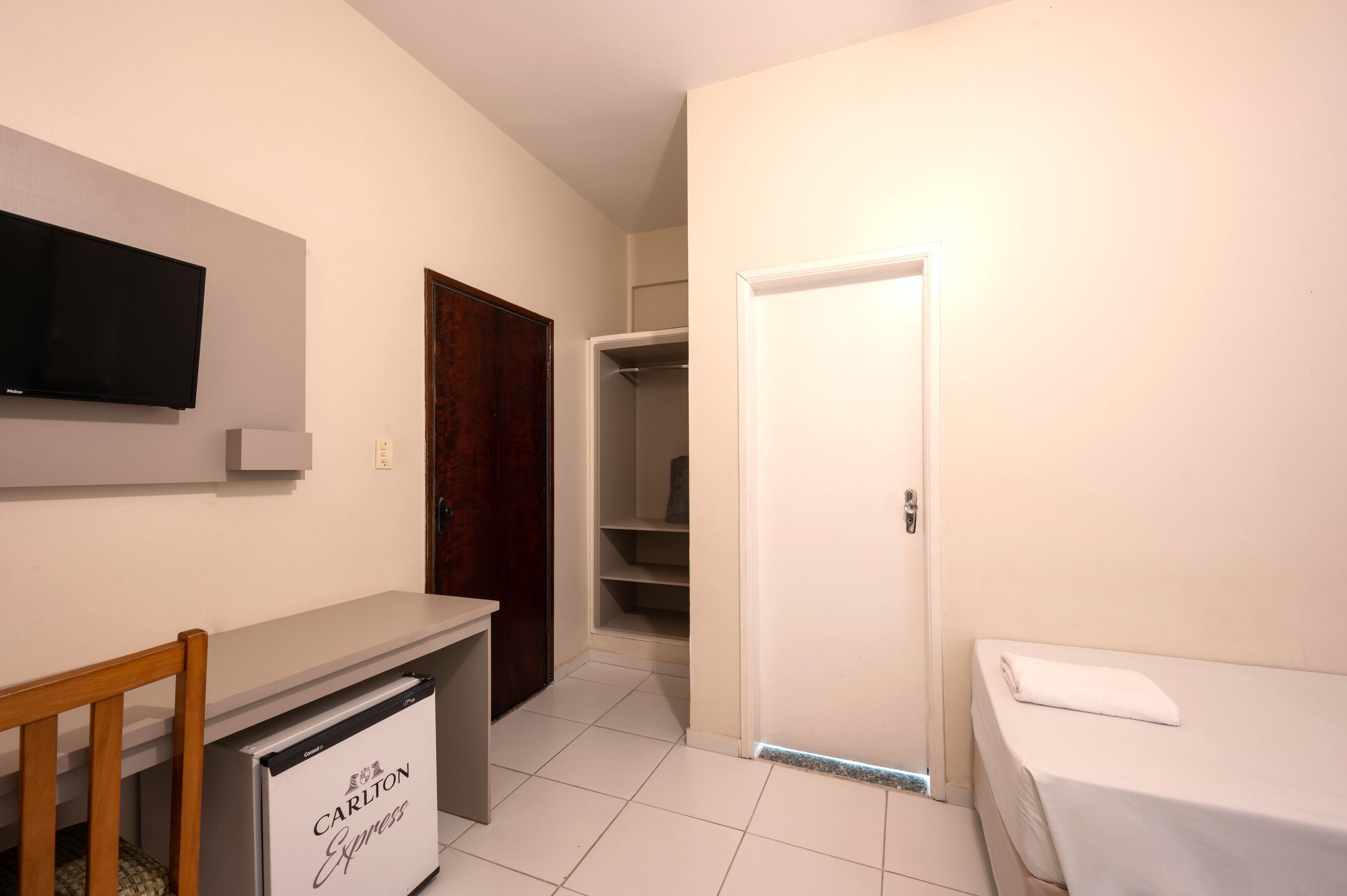 A small, clean room with a desk, chair, and a bed. There is a television mounted on the wall and a closet with shelves.