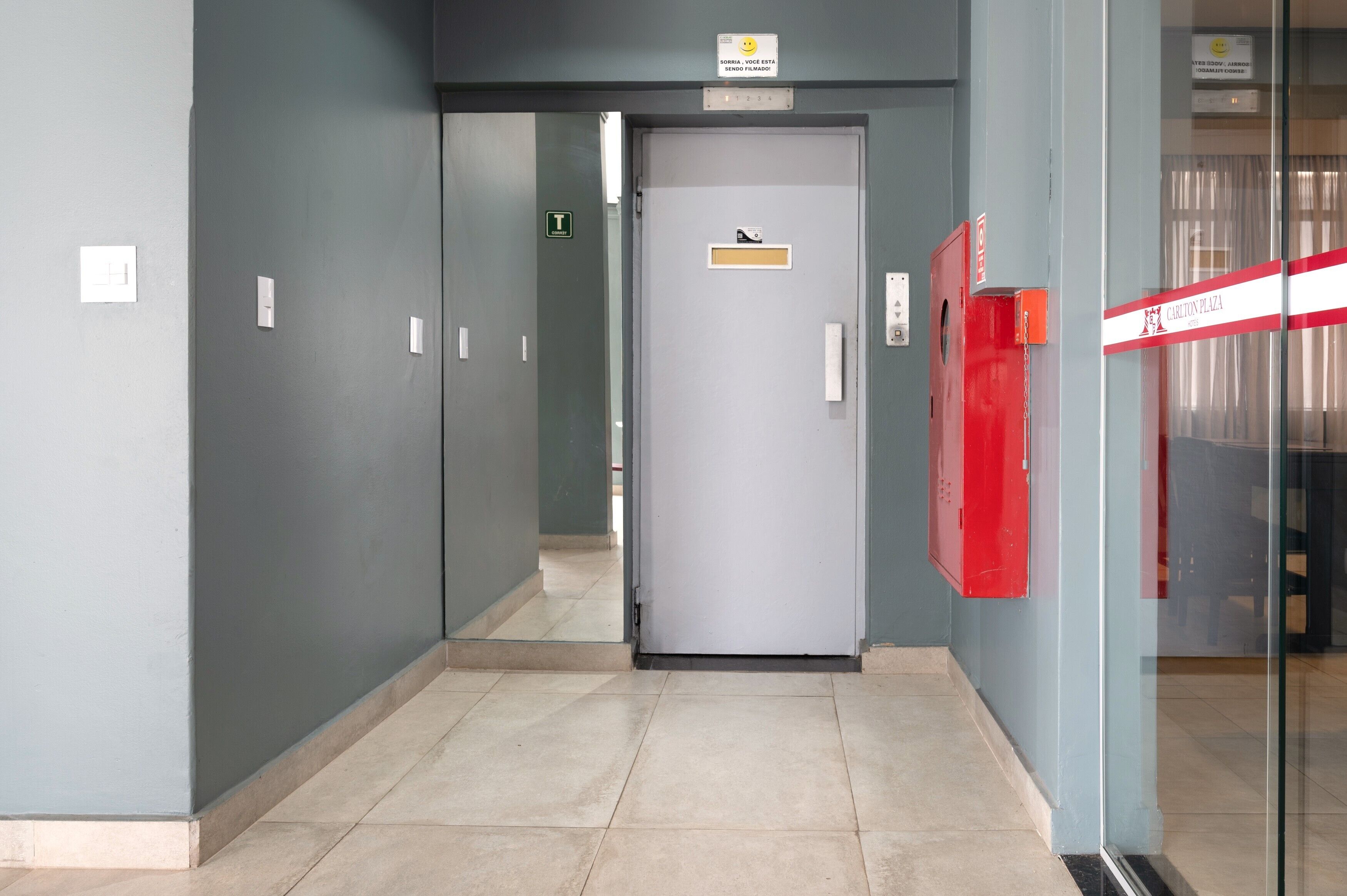 Un pasillo con una puerta gris, un extintor rojo y un letrero que indica un baño.