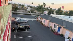 Ein Parkplatz mit mehreren Autos, ein kleines Gebäude mit einem Schild, das ein Hotel anzeigt, und im Hintergrund eine Aussicht auf Palmen und eine Straße.
