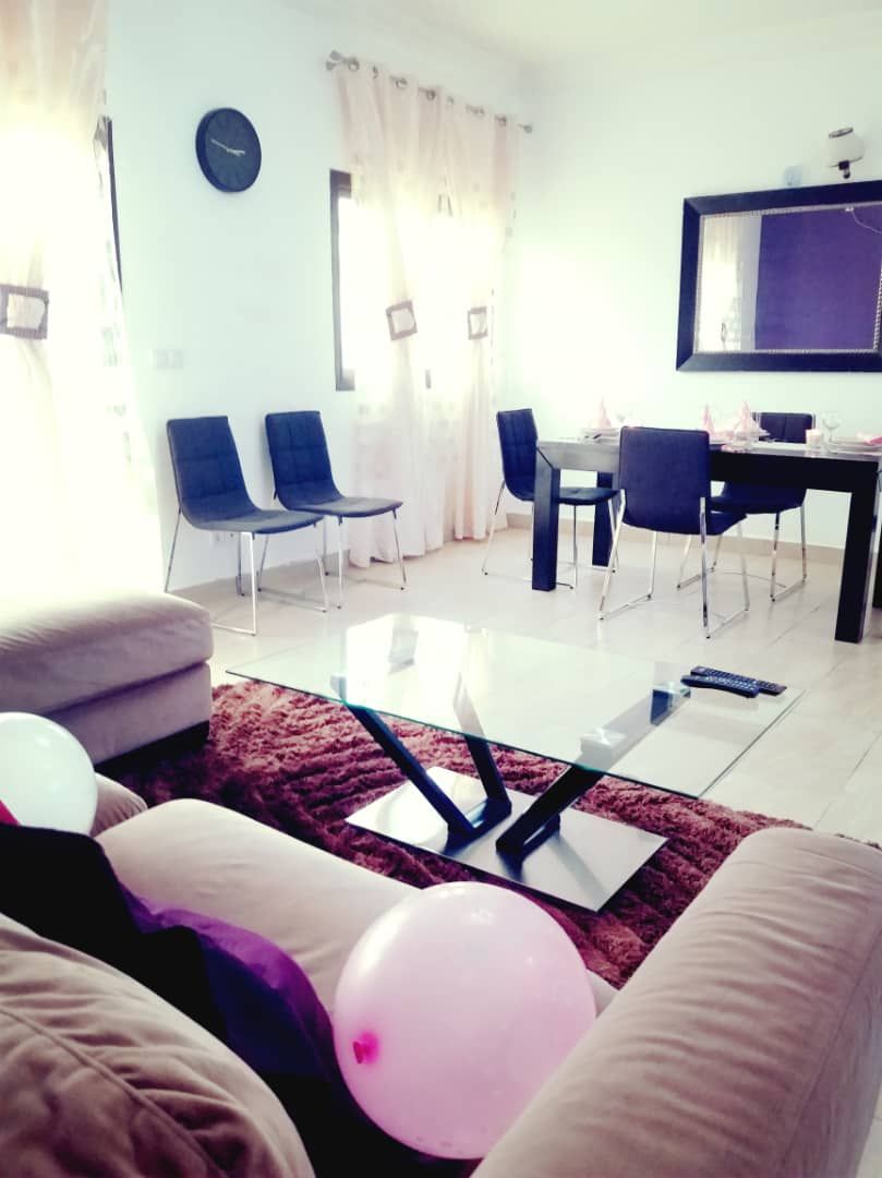 A living room with a glass coffee table, sofas, a dining table with chairs, a clock on the wall, and a flat-screen TV mounted above the table.
