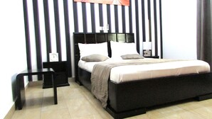 Un dormitorio moderno con cabecera negra, cama con ropa de cama blanca y gris, mesita de noche y una silla.