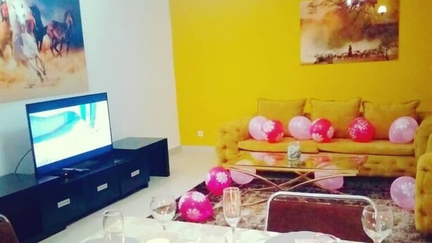 Un comedor con una mesa preparada para cuatro personas, un sofá con globos, un televisor y un aire acondicionado en la pared.