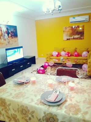 A dining area with a table set for two, a sofa in the background, and a television on a stand. - NewLand Sarl (Douala)
