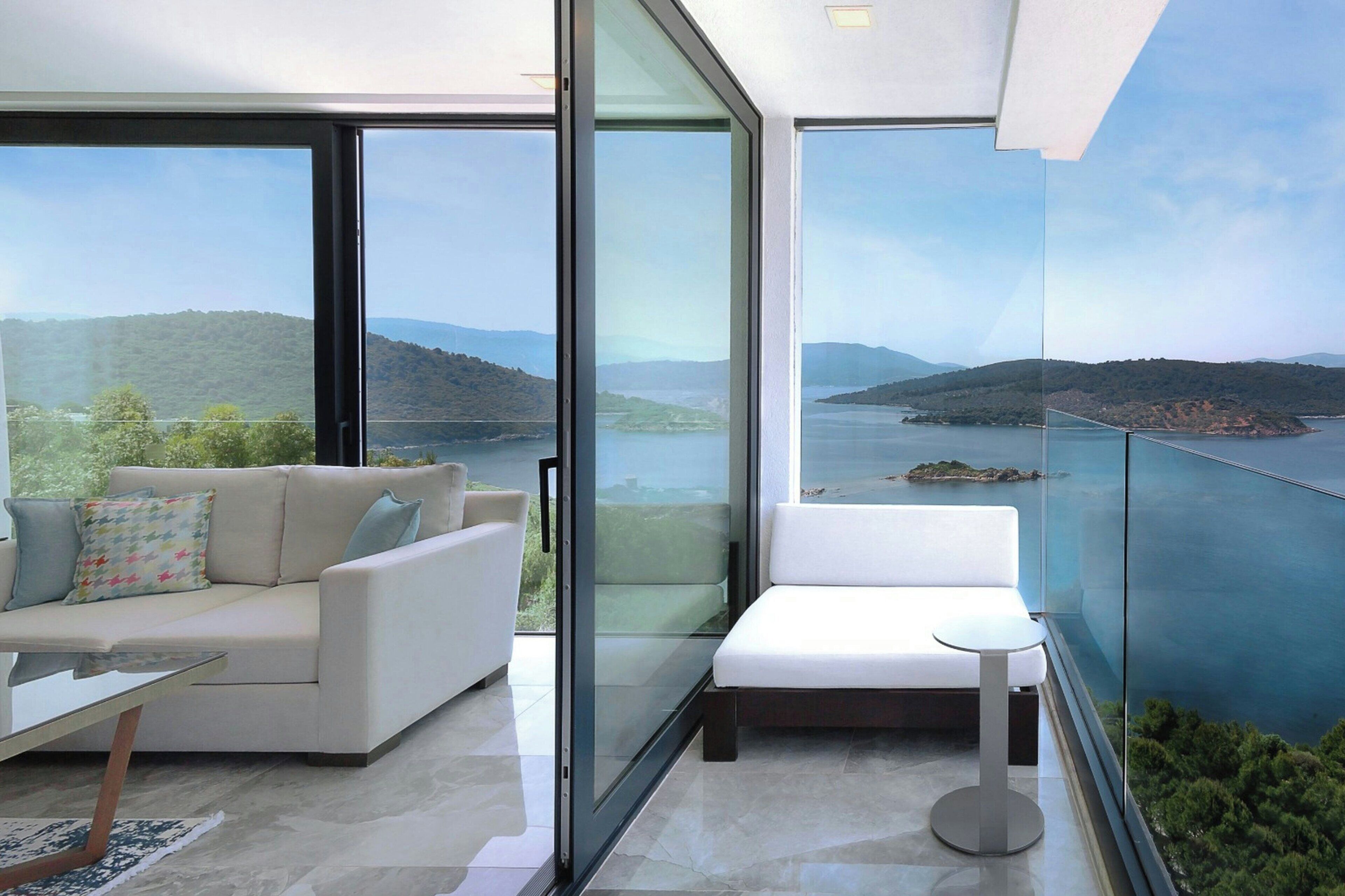A modern living room with a glass wall, a sofa, a small table, and a view of a lake and hills.