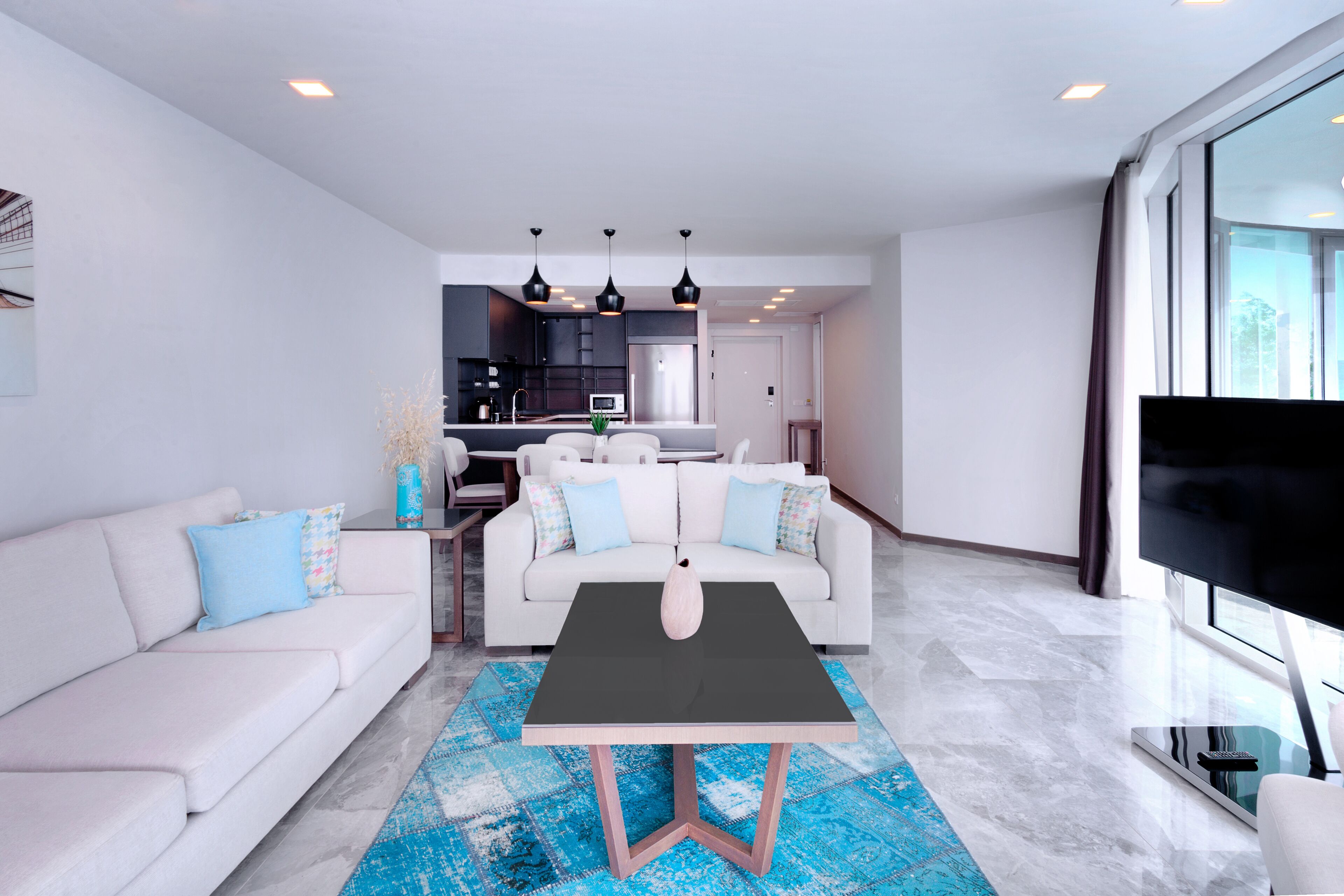 A modern living room with a white sofa, a coffee table, and a TV. There is a kitchen in the background with dark cabinets and a dining area.