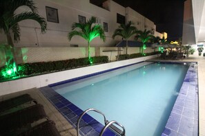 A swimming pool with a blue tiled edge, surrounded by palm trees and a white building with a brick base. - IMPERIAL TAMBAU Flat (João Pessoa)