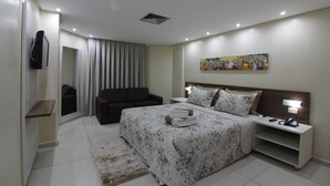 Una habitación de hotel moderna con una cama grande, una mesita de noche con lámpara, un televisor montado en la pared y un cuadro sobre la cama.