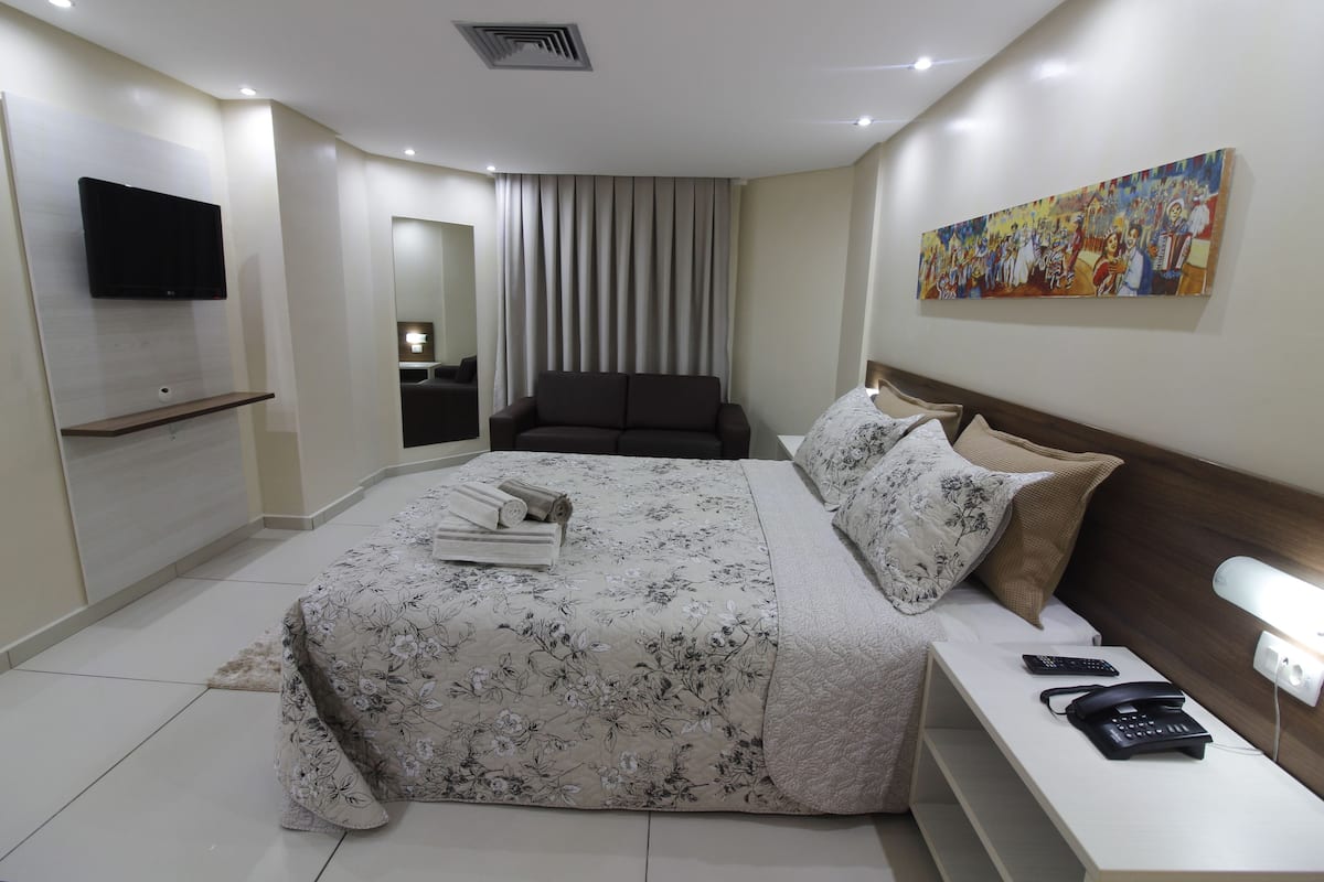 Quarto de hotel moderno com uma cama grande, televisão de tela plana, mesa de cabeceira com abajur e um quadro na parede.