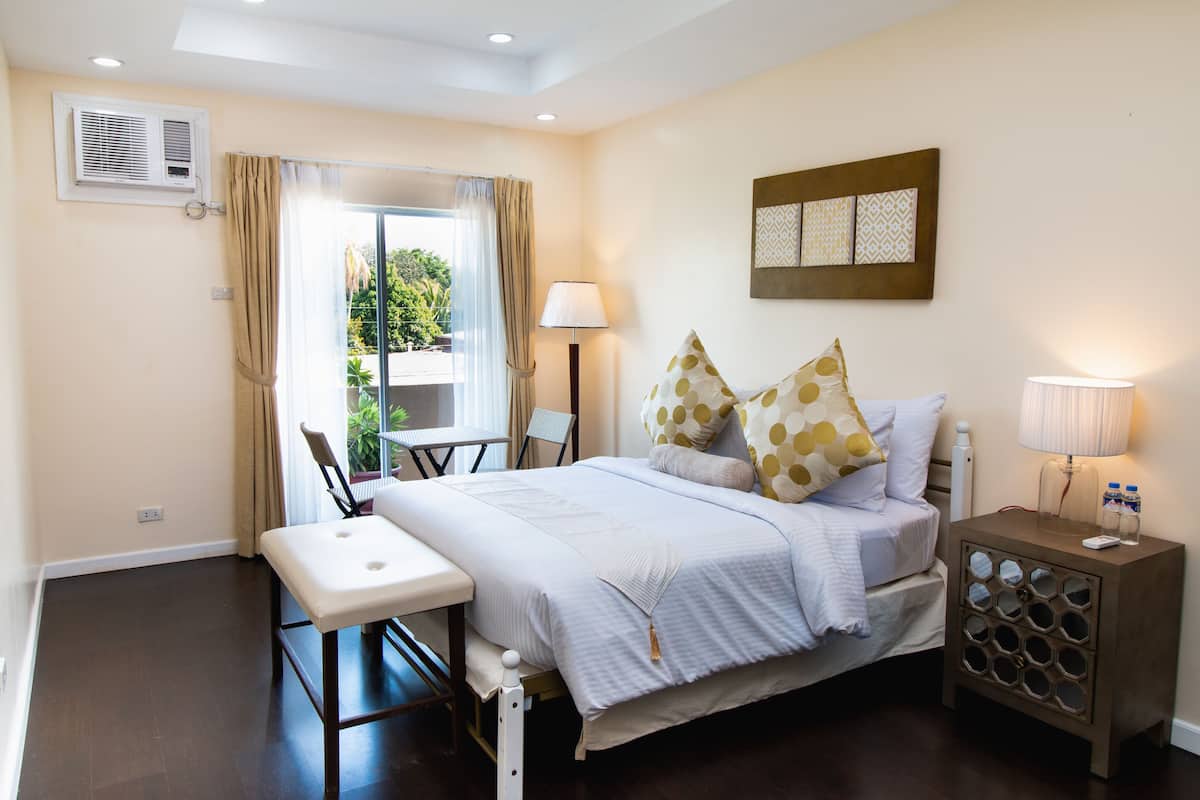 A bedroom with a bed, bedside table, lamp, and a view of greenery outside.