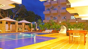 Zona de piscina con sillones de descanso y sombrillas, rodeada de plantas en macetas y un edificio de varios pisos al fondo.