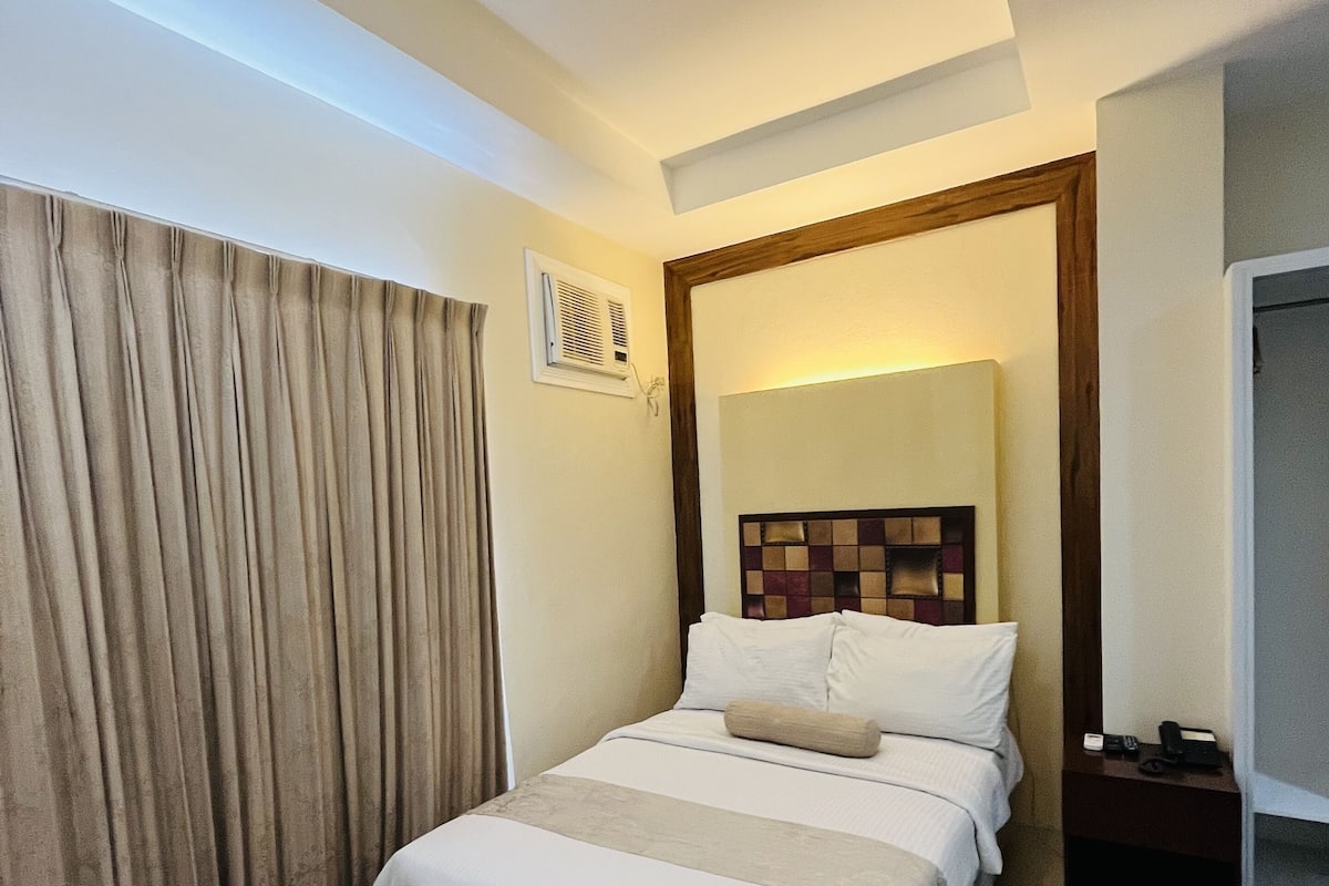 A hotel room with a bed, a nightstand, a window with curtains, and a door.