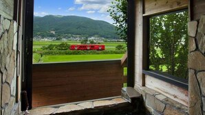 A wooden cabin with a stone exterior, a small wooden deck, and a view of a red train on tracks in the distance.
