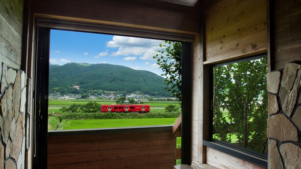 A room with a stone wall, a wooden bench, and a window overlooking a red train on tracks and green fields.