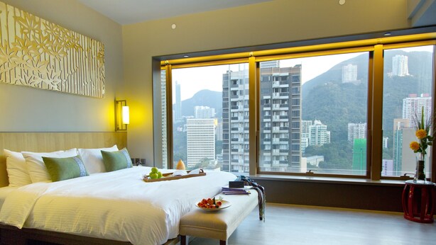 A hotel room with a large bed, a bedside table with a plate of fruit, a vase with flowers, and a view of tall buildings and mountains through large windows. - Wanchai 88 Hotel (Hong Kong)
