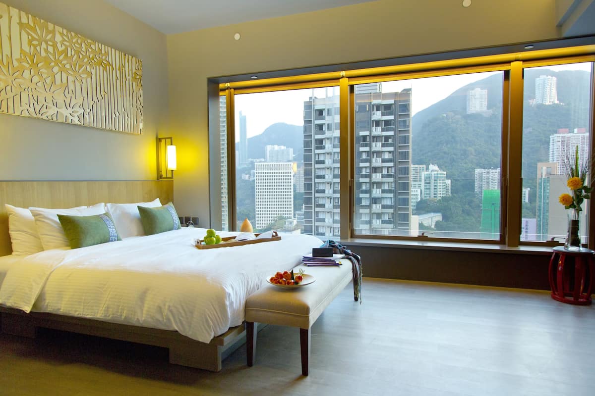 A hotel room with a large bed, a bedside table with a plate of fruit, a vase with flowers, and a view of tall buildings and mountains through large windows.