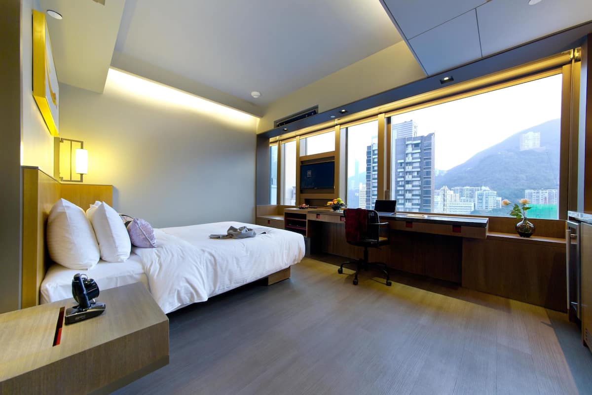 A modern hotel room with a bed, desk, and large windows offering a city view.