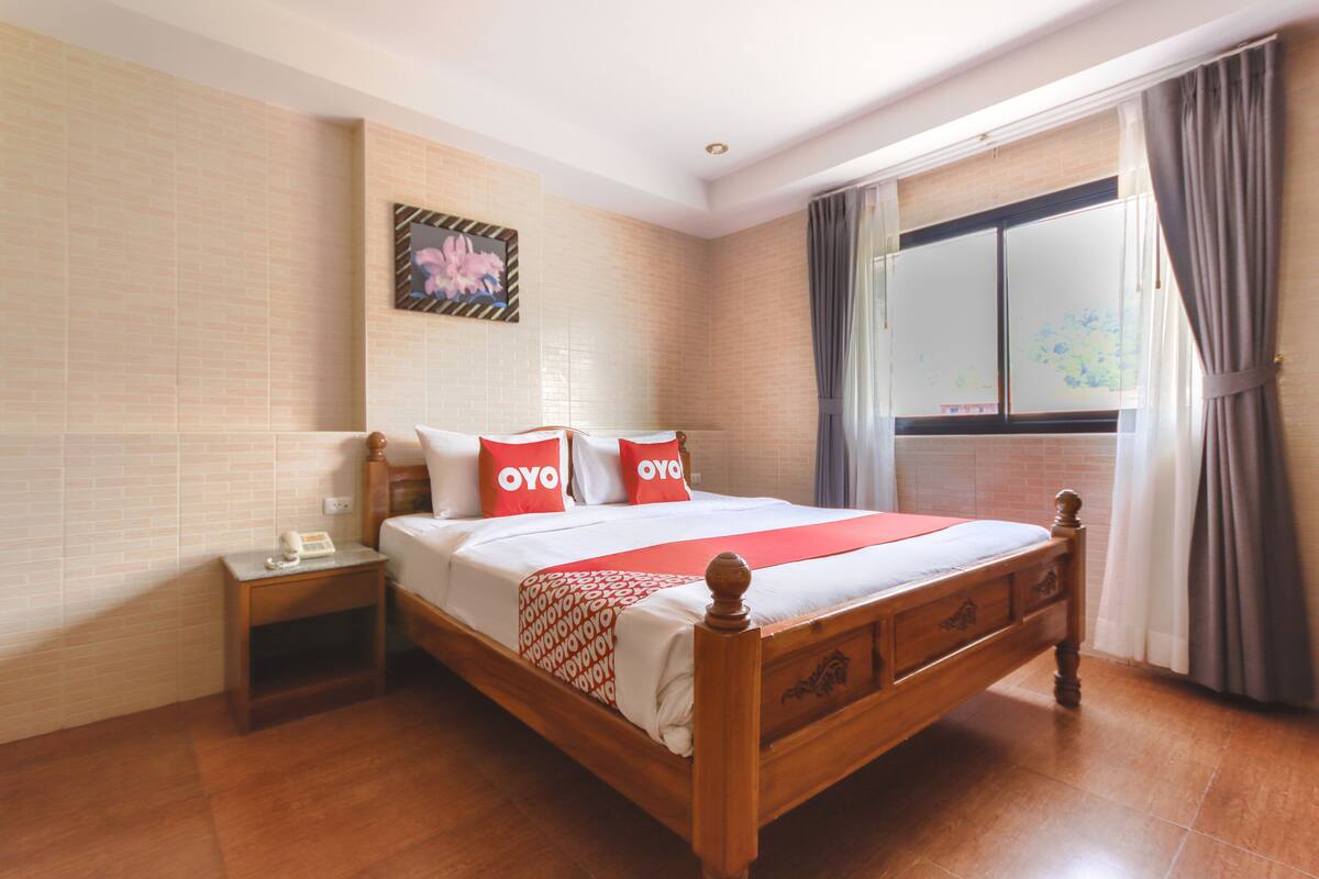 Habitación de hotel ordenada con una cama de madera, una mesita de noche, una ventana con cortinas y una obra de arte floral en la pared.