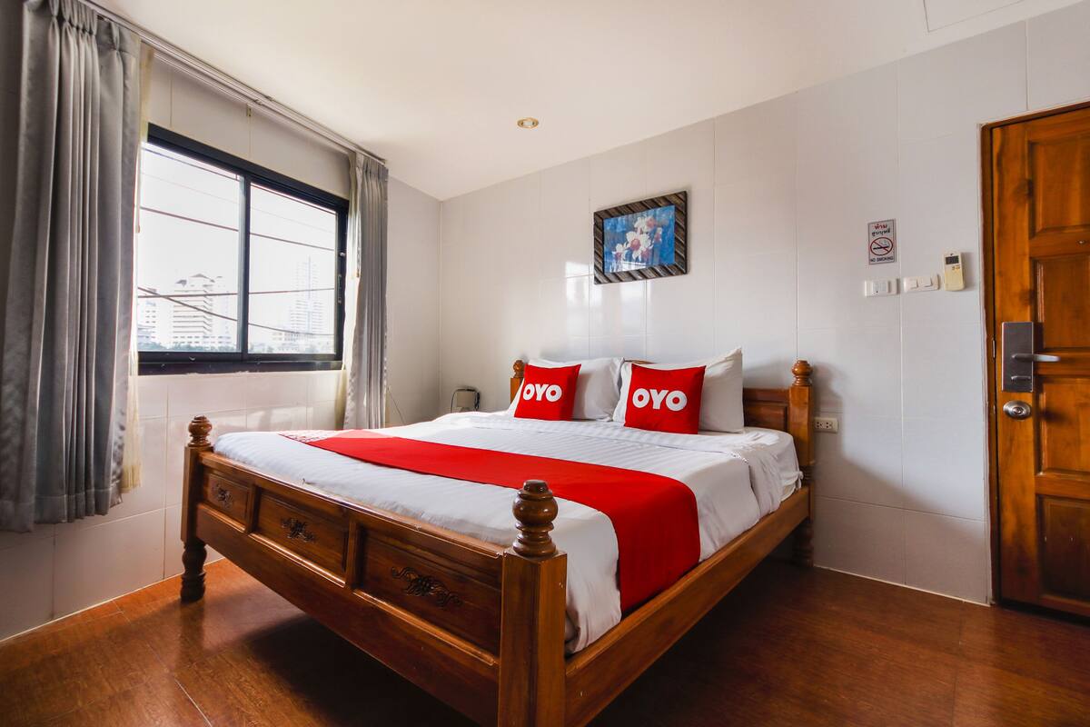 Una habitación de hotel con una cama de madera, ropa de cama blanca, almohadas rojas, una ventana con cortinas, un cuadro en la pared y una puerta.