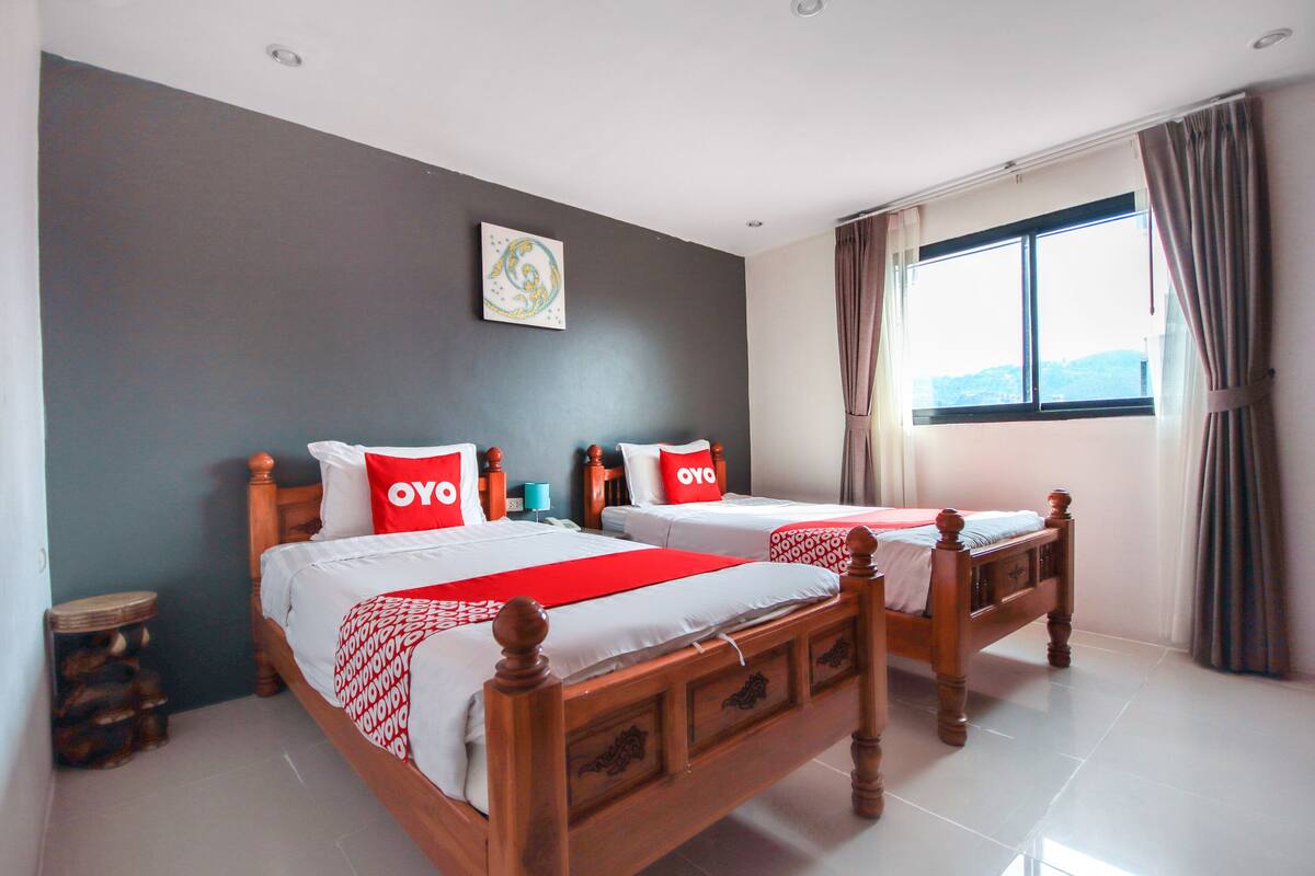 Una habitación de hotel con dos camas de madera, cada una con ropa de cama roja y blanca, una mesita de noche pequeña, una obra de arte colgada en la pared y un ventanal con cortinas.
