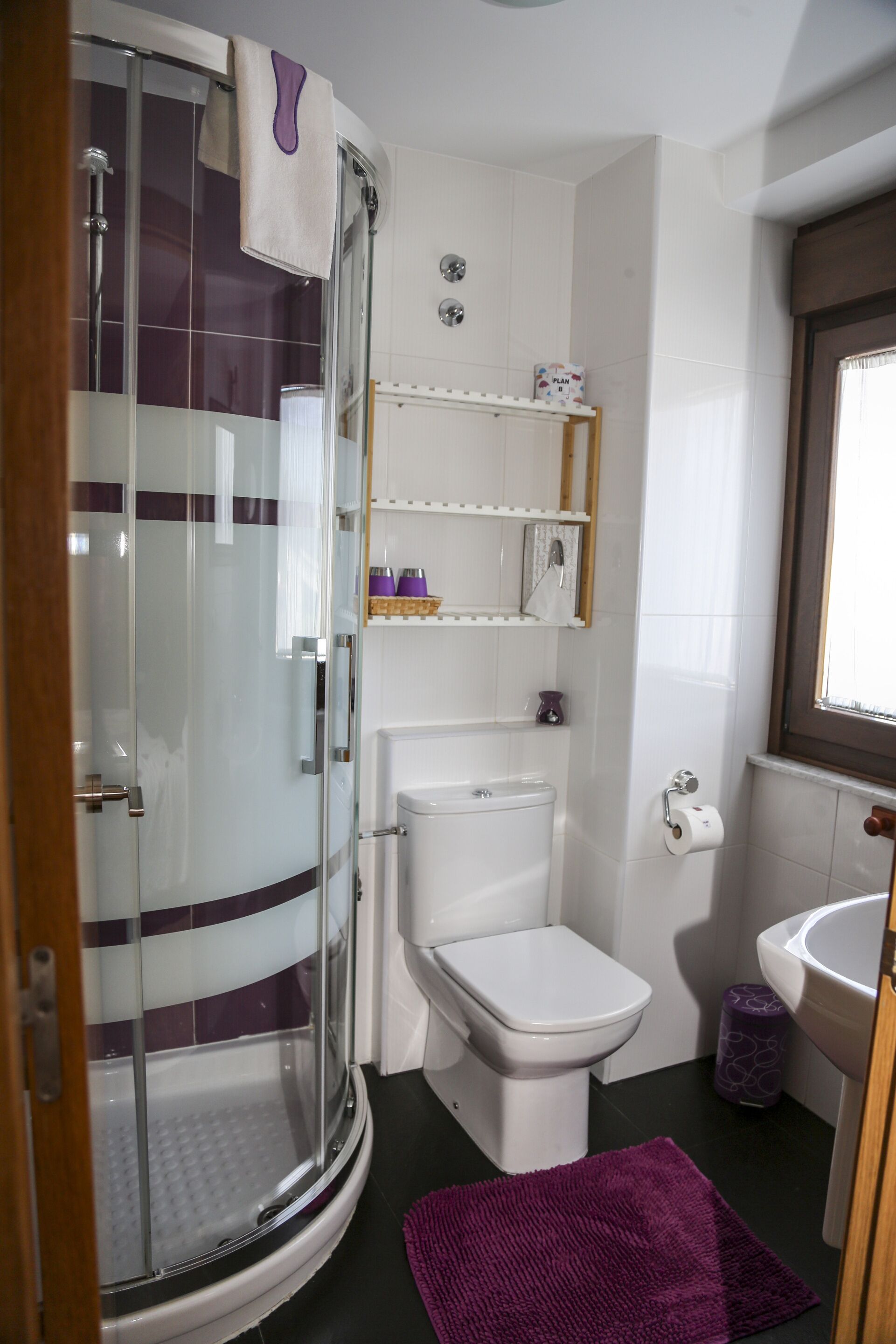 Un baño con cabina de ducha de vidrio, inodoro, lavamanos y una alfombrilla de baño morada.