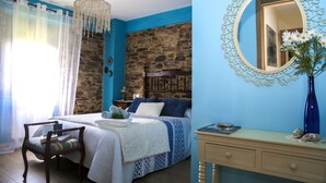Un dormitorio con pared de piedra, una cama con ropa de cama azul, una silla de madera, una mesita de noche con un jarrón y un espejo con marco decorativo.