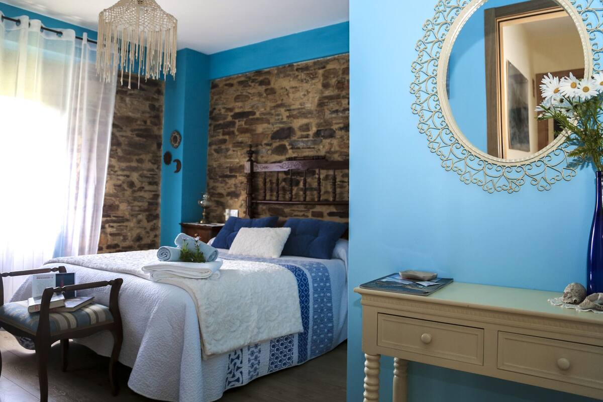 Une chambre avec un mur en pierre, un lit avec une literie bleue, une chaise en bois, une table de chevet avec un vase et un miroir avec un cadre décoratif.