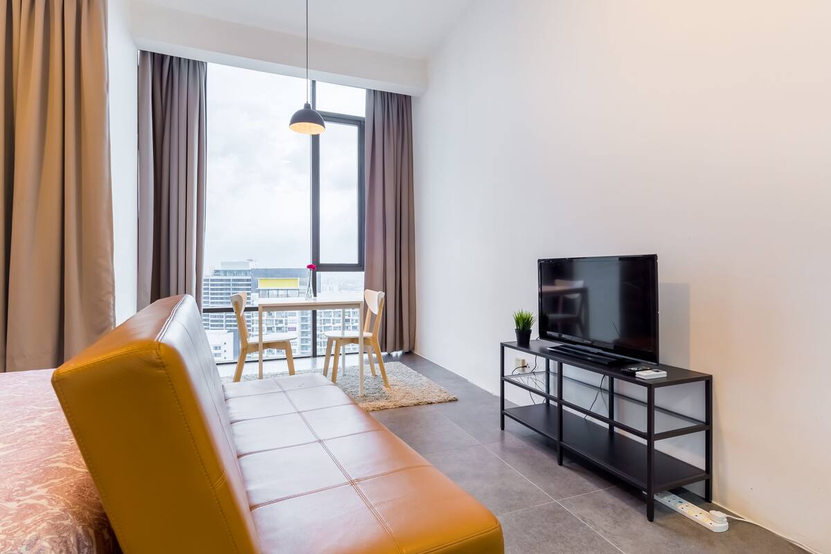 Une chambre d’hôtel moderne dotée d’une grande fenêtre, d’un canapé, d’un téléviseur posé sur un meuble noir et d’un coin repas avec des chaises.