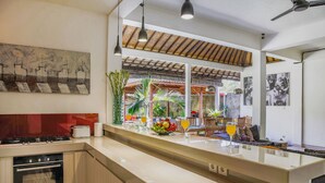 A modern kitchen with a large island, built-in oven, and a dining area with a view of a garden. - Villa Bali Caviar (Seminyak)