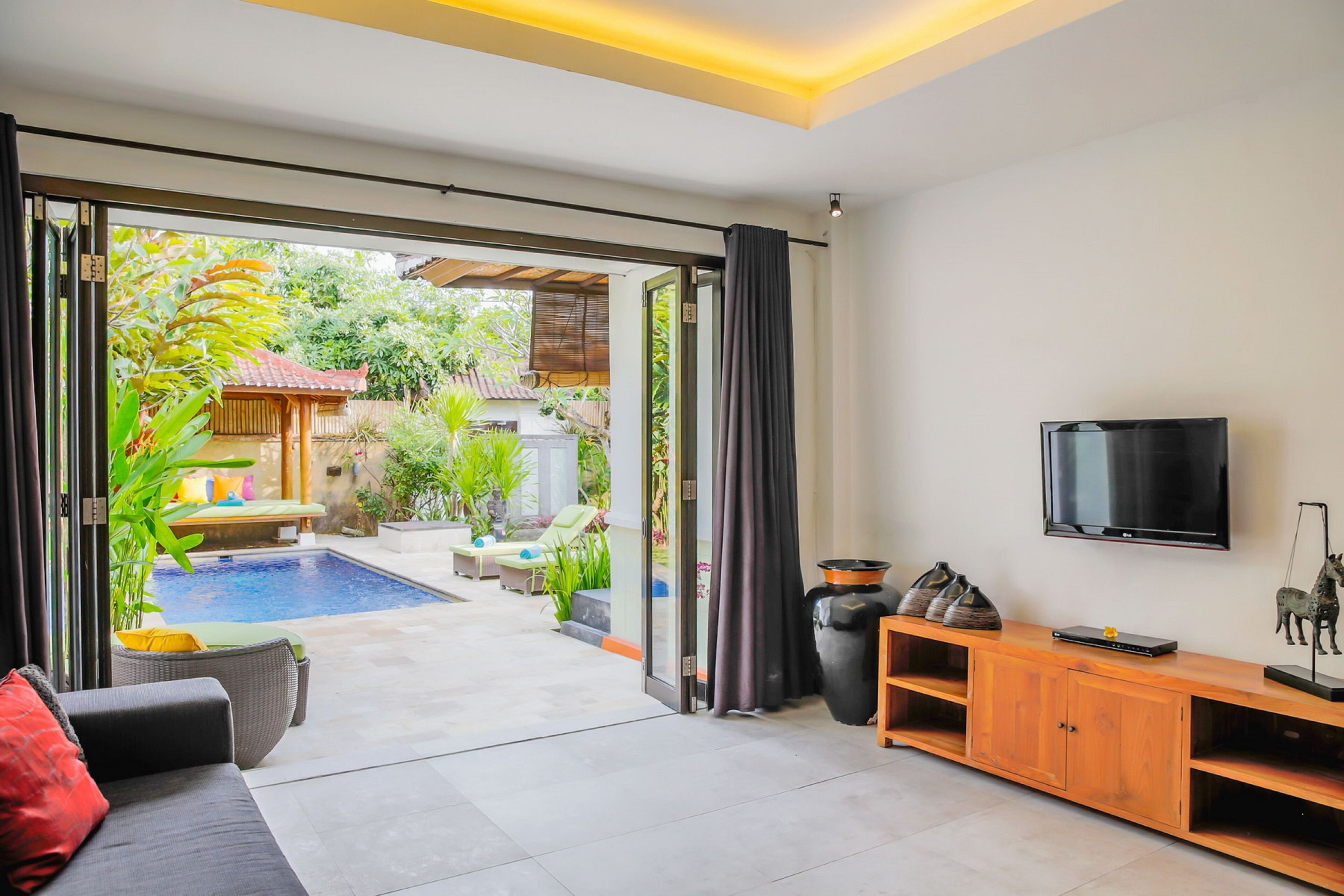 Una sala de estar moderna con televisor, un mueble de madera y un gran ventanal corredera que da a una zona de piscina.